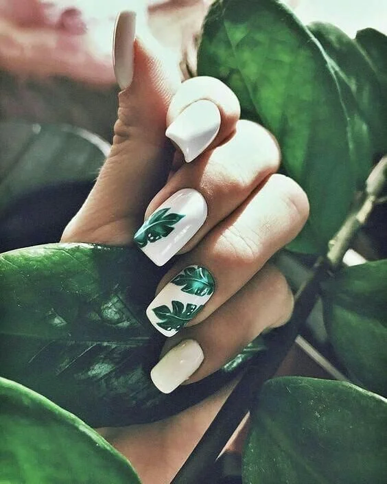 Palm Leaf Nail Art on White A close-up of a hand with white nails, two of which feature a vibrant green palm leaf design, nestled among real green leaves.